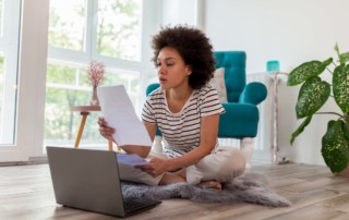 Pensive, young, black woman sitting on the floor of her home, reviewing paperwork.