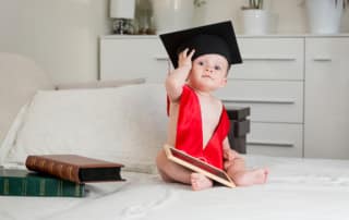 Cute baby boy in academician mortarboard sitting on bed and looking in camera