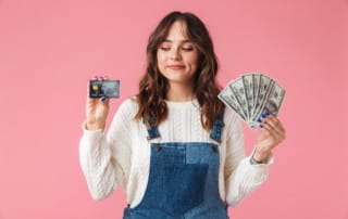 Portrait of a confident young girl holding money and showing credit card isolated over pink background