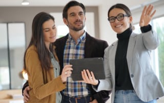 Young couple inside a house they're looking to buy, with the realtor gesturing to something in the distance