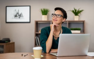 Young businesswoman sitting at a desk with her laptop, looking into the distance inquisitively