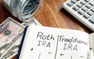 An open journal on a table with the words Roth IRA and Traditional IRA written in marker at the top. Calculator and money in the background.