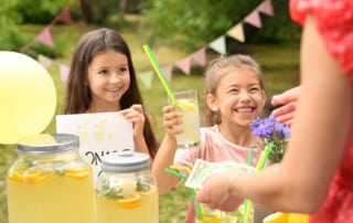 Two young girls running a lemonade stand on a sunny day, smiling as they hand someone a drink.