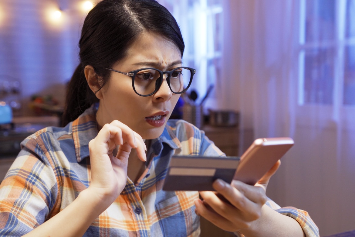 Dark-haired woman with glasses, in her 30s, looking at her phone in shock as she holds a credit or debit card.