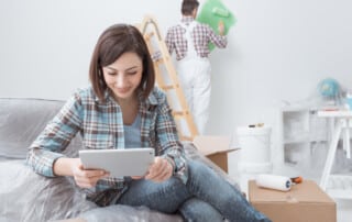 Young woman looking down at her tablet while a painter paints a wall green in the background.