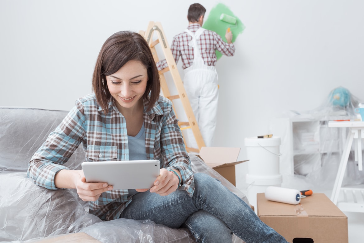 Young woman looking down at her tablet while a painter paints a wall green in the background.